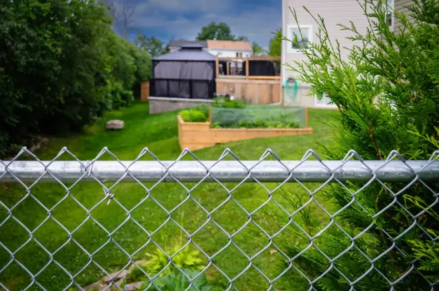 Close-up of galvanized chain link mesh and top rail with a backyard, raised garden beds, and a house behind it