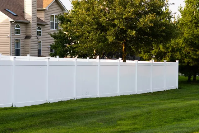 White 6-foot vinyl privacy fence with capped posts running along a lawn beside a two-story house and a large shade tree — a typical PVC privacy run